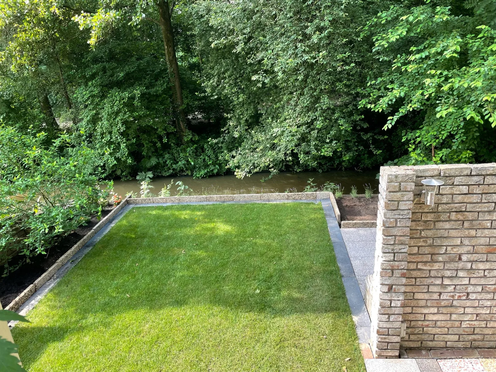 Tuin met gras en uitzicht op een beek omringd door bomen.